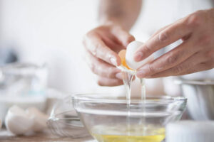 Ręce rozbijające jajko nad miseczką, aby pokazać co można zrobić z białek na szybko po przygotowaniu pączków lub ciast.